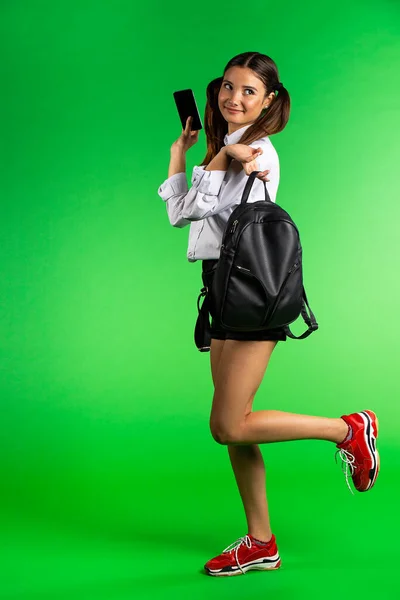 Молодая красивая женщина в коротком платье позирует с рюкзаком и смартфоном на зеленом фоне с копировальным пространством — стоковое фото