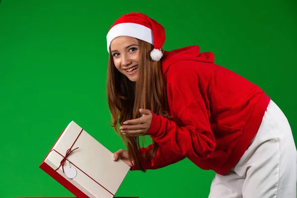 Blank glimlachen brunette vrouw in santa kleding kijkt naar camera met een kerstcadeau, geïsoleerde groene achtergrond — Stockfoto