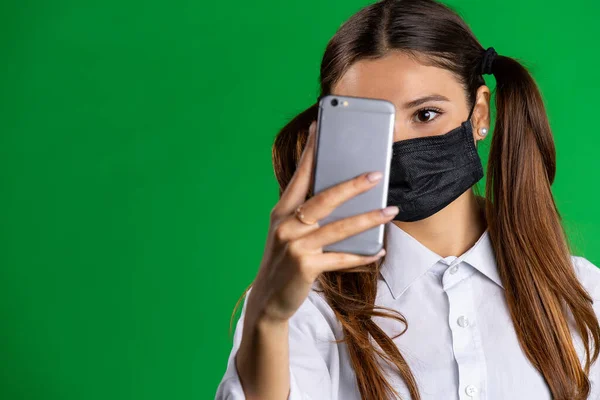 Nastoletnia uczennica w masce robi selfie na smartfonie. Współczesna nastolatka z warkoczami ubrana w luźne ubrania i czarną maskę ochronną robiąca selfie na telefonie komórkowym na zielonym tle — Zdjęcie stockowe