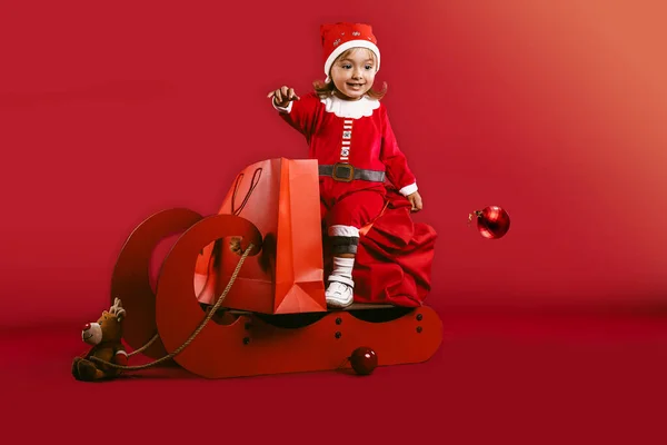 Feliz Navidad y Feliz Año Nuevo. A la niña le gusta montar en trineo por decoraciones de Navidad con juguete de renos y paquete de compras de Navidad. Diversión invernal. Niña divertirse Nochebuena sobre fondo rojo — Foto de Stock