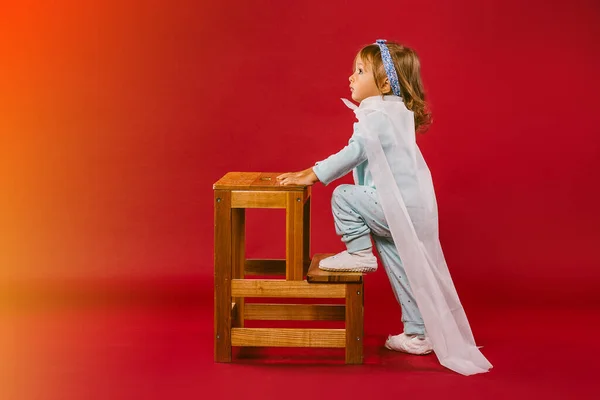 Parece mágico. Una niña pequeña vestida en pijama y con una capa de superhéroe hecha a mano blanca, sube la escalera de madera mirando hacia arriba.Aislada sobre fondo naranja a rojo con espacio lateral libre — Foto de Stock