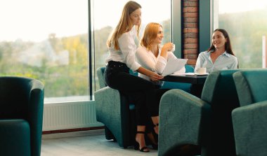 Arkadaşlar modern ofiste kahve içerek eğleniyorlar. Yeni proje altında beyin fırtınası yapıyorlar. Genç kadınlar kahve molasında birlikte oturmayı tartışırlar..