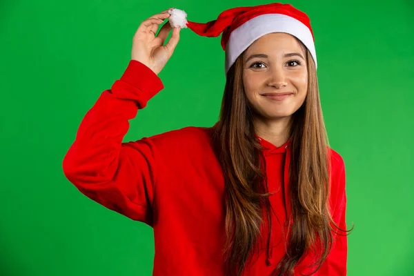 Femme brune au chapeau de Père Noël et pull rouge souriant, regardant la caméra sur fond vert isolé — Photo