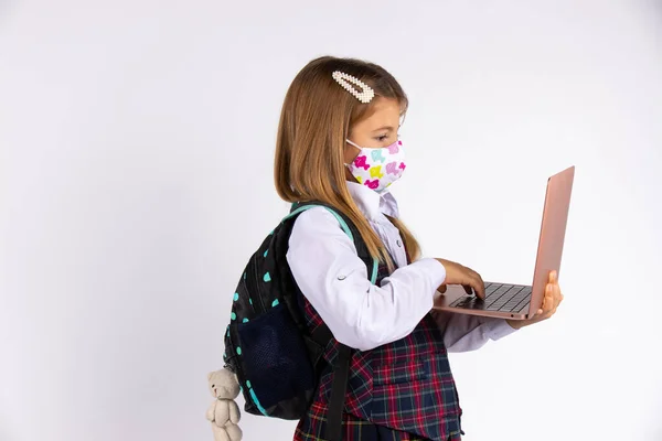 Retrato de colegiala de primaria en uniforme con máscara médica facial que regresa a la escuela después de la cuarentena y el bloqueo covid-19. Mochila y computadora portátil en las manos. — Foto de Stock