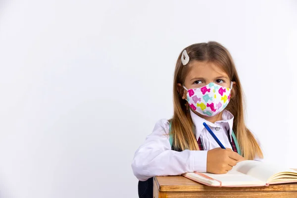 Niña en la escuela disgustada con una máscara protectora sentada en la mesa en la lección. Nuevas reglas escolares normales, salud y concepto de epidemia — Foto de Stock