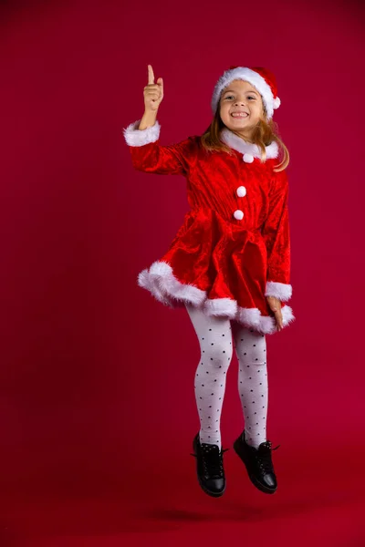 Colegiala divertida en traje de Navidad con sombrero de Santas saltando sobre fondo de color rojo con mucho espacio. Kid apunta hacia arriba. — Foto de Stock