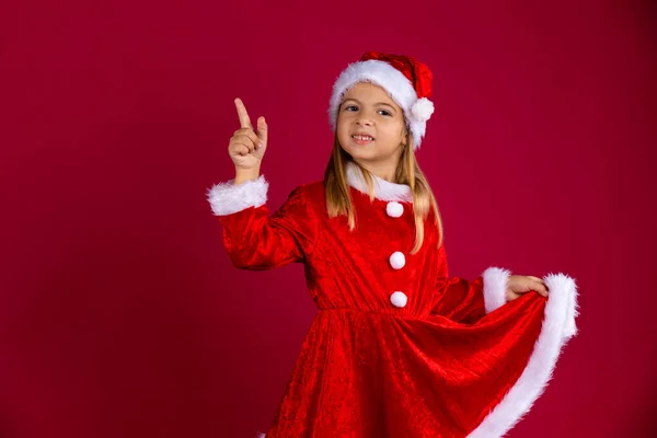 Hermosa niña con traje de Santa Claus y sombrero de invierno. Retrato de un niño sosteniendo el vestido rojo a mano y apuntando hacia el dedo. Aislado sobre fondo rojo con espacio libre — Foto de Stock