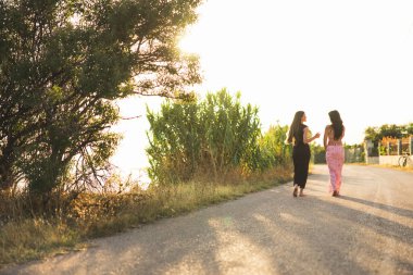 İki arkadaş, gün batımında manzaralı bir sahil yolunda birlikte sakin bir gezinti yapmaktan zevk alır.