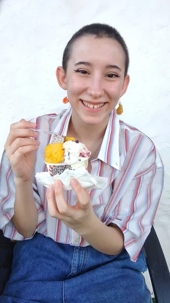 Dondurma yiyen genç bir delikanlı. Gülümsemesi ve mutluluk ifadesi var. Üzerinde kırmızı çizgili beyaz gömlek, kot pantolon ve küpe var.