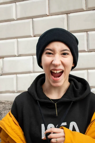 Mutluluk ifadesiyle çığlık atan genç bir kız. Siyah yün şapka, siyah kazak ve sarı palto giyen bir fayans duvarının önünde..