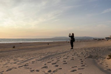 Lizbon şehir merkezine yakın Costa da Caparica sahilinde yürüyen güzel beyaz kadın. Caparica 'da gün batımında plajda dinlenen deri ceketli güzel bir kadın. Portekiz, 2020