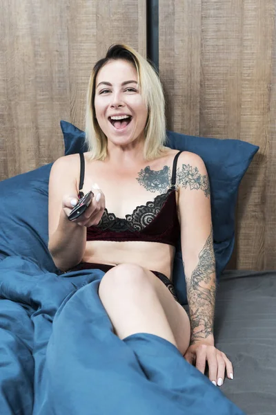 Portrait of a laughing sexy woman with tattoos with a remote control in her hand on the bed