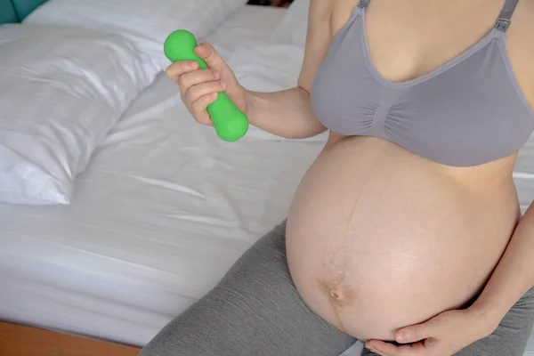 Молода вагітна жінка тримає гантелі на ліжку, концепція здорового — стокове фото