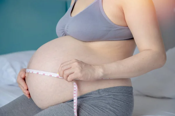 Young pregnant woman measuring tummy with measuring tape on bed — Stock Photo, Image