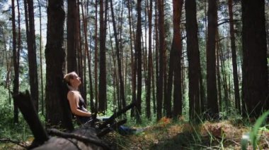 Ormandaki bir kız ağaca yaslanmış, kulaklıklarıyla müzik dinliyor. Doğada dinlenmek. Arka planda bulanık ağaç siluetleri var. Dijital detoks. Akıl sağlığı. Doğa sesi
