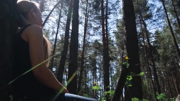森林里的一个女孩靠着树坐着 耳机在听音乐 在大自然中休息 背景中模糊了树木的轮廓 数字排毒 精神健康 大自然的声音 — 图库视频影像