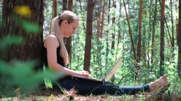 森林里年轻漂亮的女人靠在树上 在笔记本电脑上工作 在大自然中休息 背景中模糊了树木的轮廓 数字排毒 精神健康 大自然的声音 — 图库视频影像