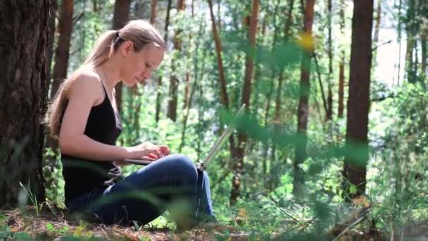 森の中の若い可愛い女性たちは木に寄りかかってノートパソコンで働いています 自然の中で休みなさい 背景には木々のシルエットがぼやけていました デジタルデトックス 精神衛生だ 自然界の音 — ストック動画