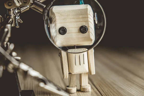 wooden robot android stands on a table under a magnifying glass. metal clamps with a magnifying glass. the face of the robot is enlarged in a magnifying glass. mechanism assembly concept