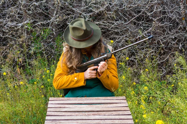 Blonde scout girl in green vest and green hat preparing weapon for hunting.