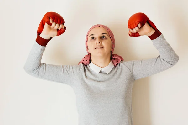 Portrait of a woman with cancer and depression locked in her home thinking. Pink headscarf and red boxing gloves. Fighting against cancer.
