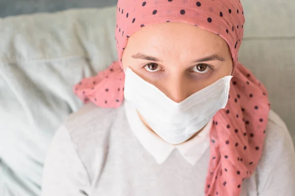 Portrait of a woman with cancer and depression locked in her home thinking. Pink headscarf and face mask. Quarantine coronavirus.