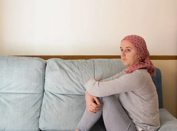 Portrait of a woman with cancer and depression locked in her home thinking. Sitting on the sofa. Pink headscarf. Quarantine coronavirus.