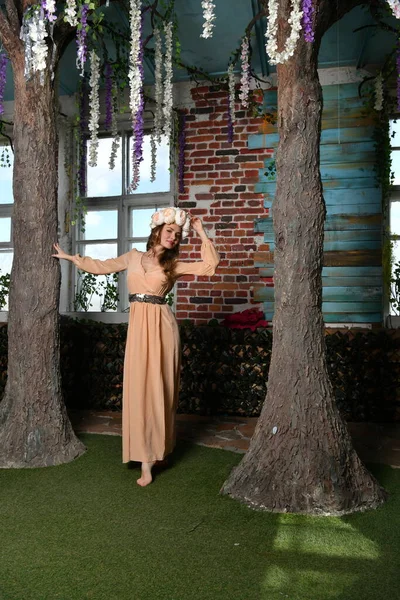 Bej elbiseli bahar kızı ve kafasında çiçek çelengi çiçek ve ağaçların arkasında poz veriyor.