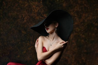 Siyah şapkalı ve bordo bir gece elbisesi giymiş güzel bir kız kendini kollarına sarmış.