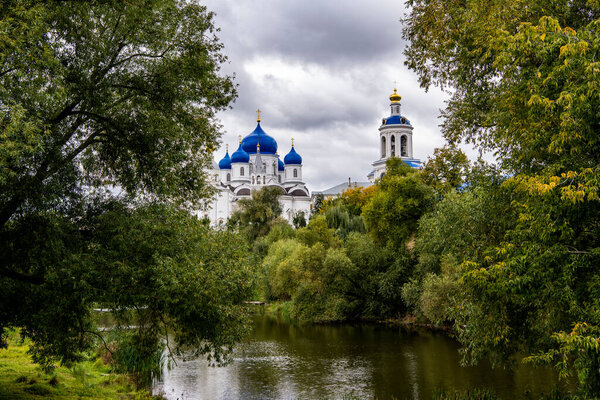 старый белый монастырь с голубыми куполами на фоне зеленых деревьев и реки