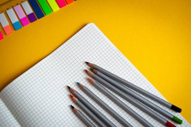 Sarı arkaplanda kalemler, keçeli kalemler ve defterler var.