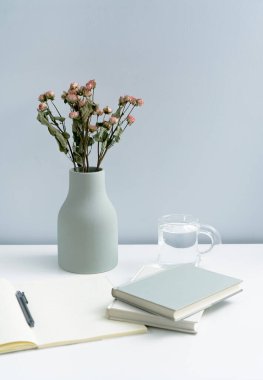 Vazo ve defter. Beyaz masanın üzerinde kurumuş çiçekler var.