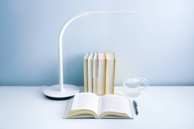 Açık bir kitap beyaz bir masanın üzerine lambayla konur. Düzenlenmiş bir yığın kitap ve beyaz bir masada şeffaf bir bardak.