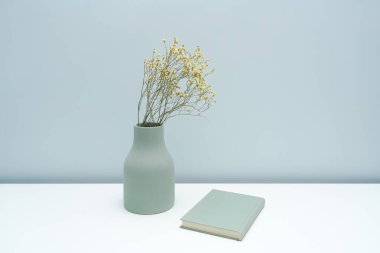 Vazo ve defter. Beyaz masanın üzerinde kurumuş çiçekler var.