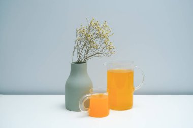 Kurumuş çiçekli bir vazo ve beyaz masada bir bardak portakal suyu..