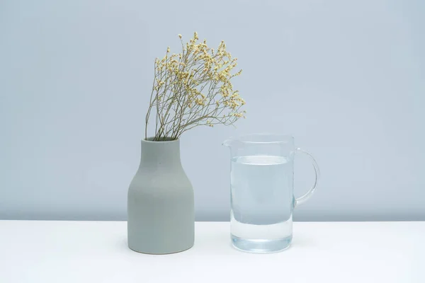 A vase with dried flowers and a glass of water on the white table.