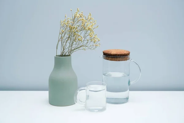 A vase with dried flowers and a glass of water on the white table.