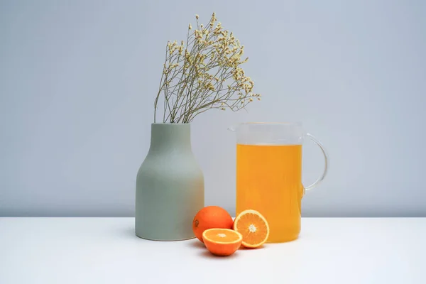 A vase with dried flowers and a glass of orange juice on the white table.