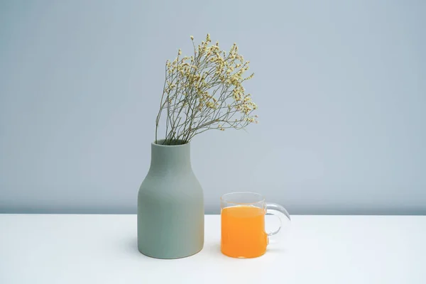 A vase with dried flowers and a glass of orange juice on the white table.