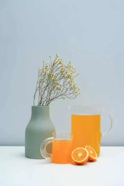 A vase with dried flowers and a glass of orange juice on the white table.