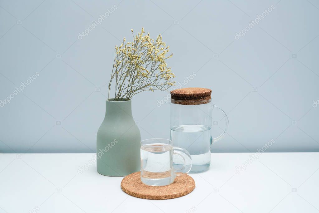 A vase with dried flowers and a glass of water on the white table.