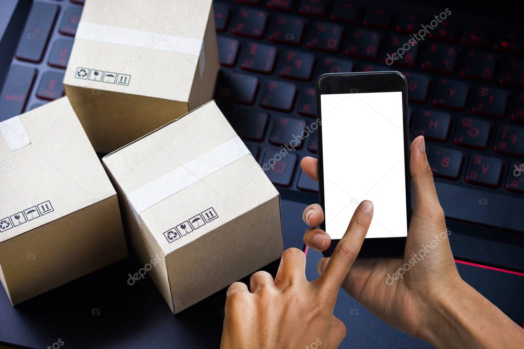 Hand holding smartphone white screen mock up.Online shopping concept e-commerce delivery buying service. square cartons shopping on laptop keyboard, Using smartphone to confirm order via the internet.