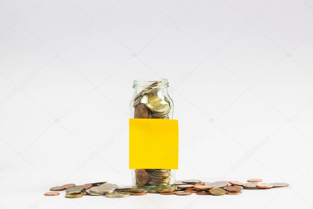 Savings, Investment growing money concept : A full coins in a clear glass jar with a empty yellow post paper and a coin on the floor on white background.