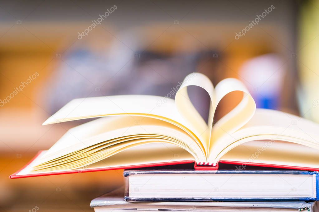 Love to read a book concept : Open book with heart shape page on stack book wood table and blurred bookshelf. Education background, back to school concept.