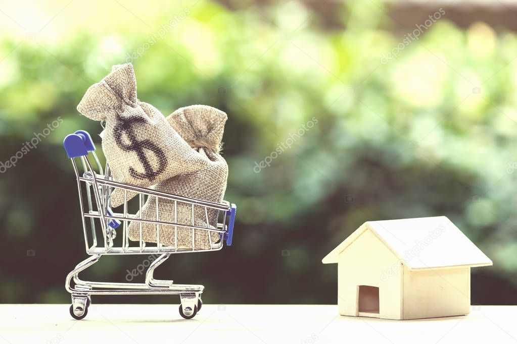 Home loan, mortgages, debt, savings money for home buying concept : US dollar money bag in shopping cart, residential, house on table against green nature background. Exchange of finances and houses.