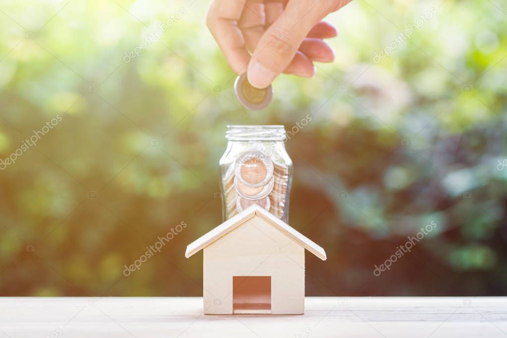Saving money, home loan, mortgage, a property investment for future concept : A man hand putting coins over small residence house and glass jar with green nature background. A sustainable investment.