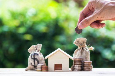 Saving money, home loan, mortgage, a property investment for future concept : A man hand putting money coin over small residence house and money bag clipart