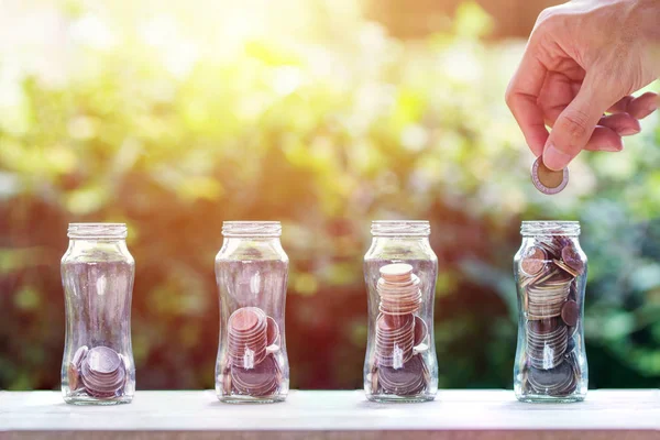 Financial investment / Savings money growing concept : Hand holding coin over four step coins in glass jar on wood table with nature background. Conceptual saving money for growing business and future
