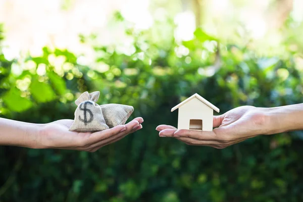 Home loan, lending, mortgage, transforming assets into cash concept : Hand holding home model change to money with green nature as background. Conceptual changing residence into cash.