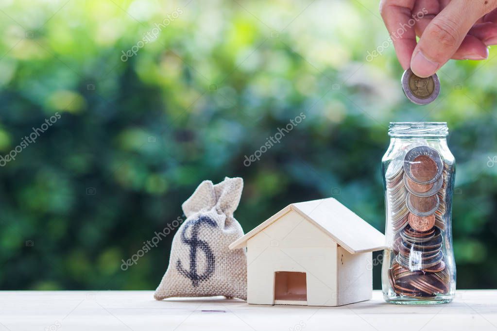 Home loan, mortgages, debt, savings money for home buying concept : Hand holding coin over glass jar. US dollar in a money bag, small residential, house model on table against green nature background.